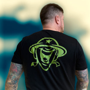 Man wearing a black t-shirt with a green happy soldier graphic on the back, standing against a blurred natural background.