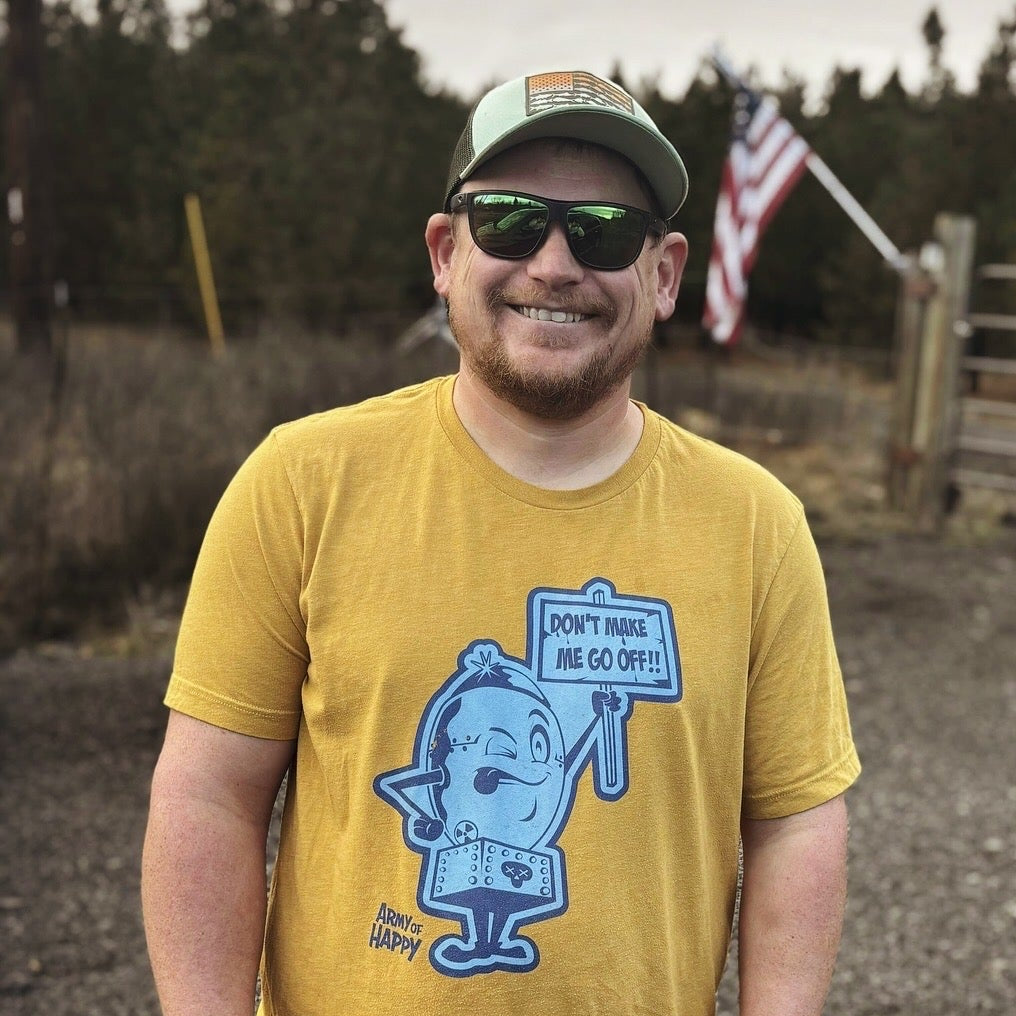Man wearing a yellow Army of Happy "Atom Bomb" t-shirt with a graphic design and text saying "Don't Make Me Go Off!" , standing outdoors with trees and an American flag in the background.