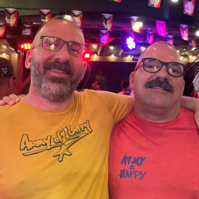 Two men wearing colorful t-shirts with Army of Happy text, posing together in a lively indoor setting.