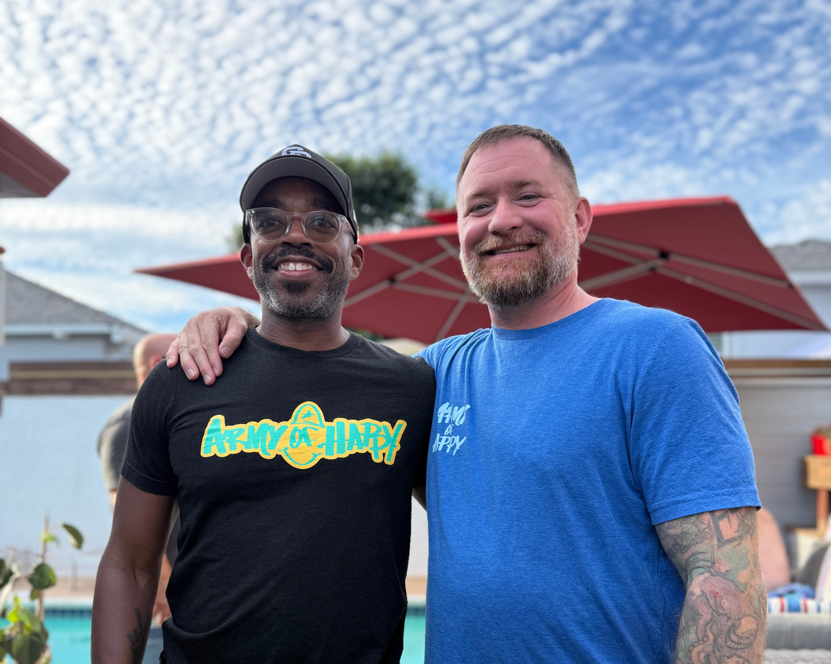 Two men standing together outdoors by a pool, smiling. The man on the left wears a black t-shirt with a colorful 'Army Jolly' graphic and a black cap.